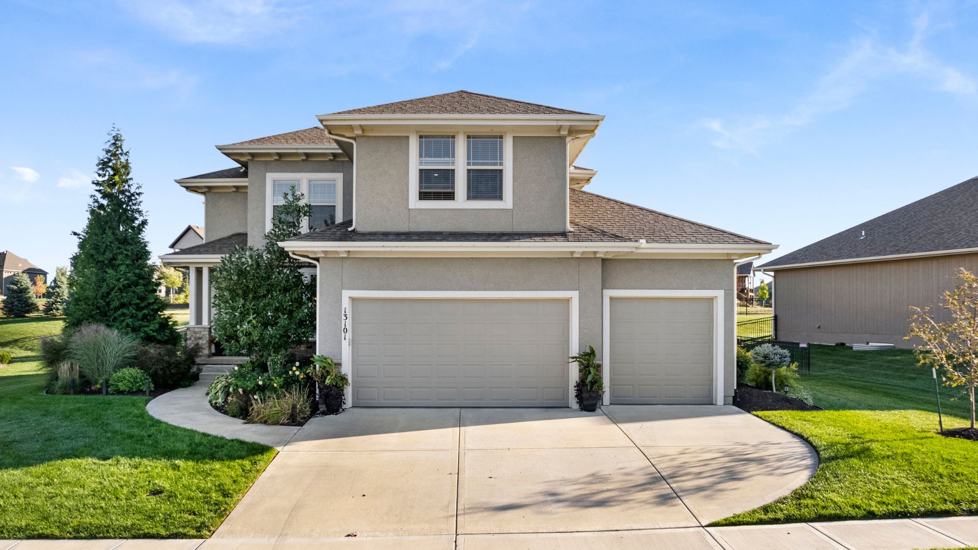 Front photo of home that has gray stucco, large driveway and luscious landscaping.