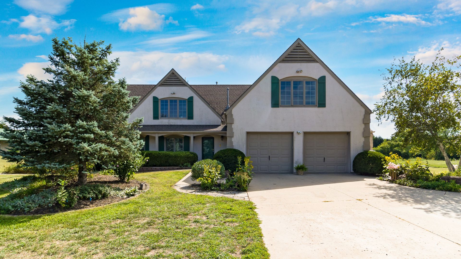 Front photo of a tan home with gray garage doors and green shutters surrounded by trees and landscaping. Large driving leading to home.