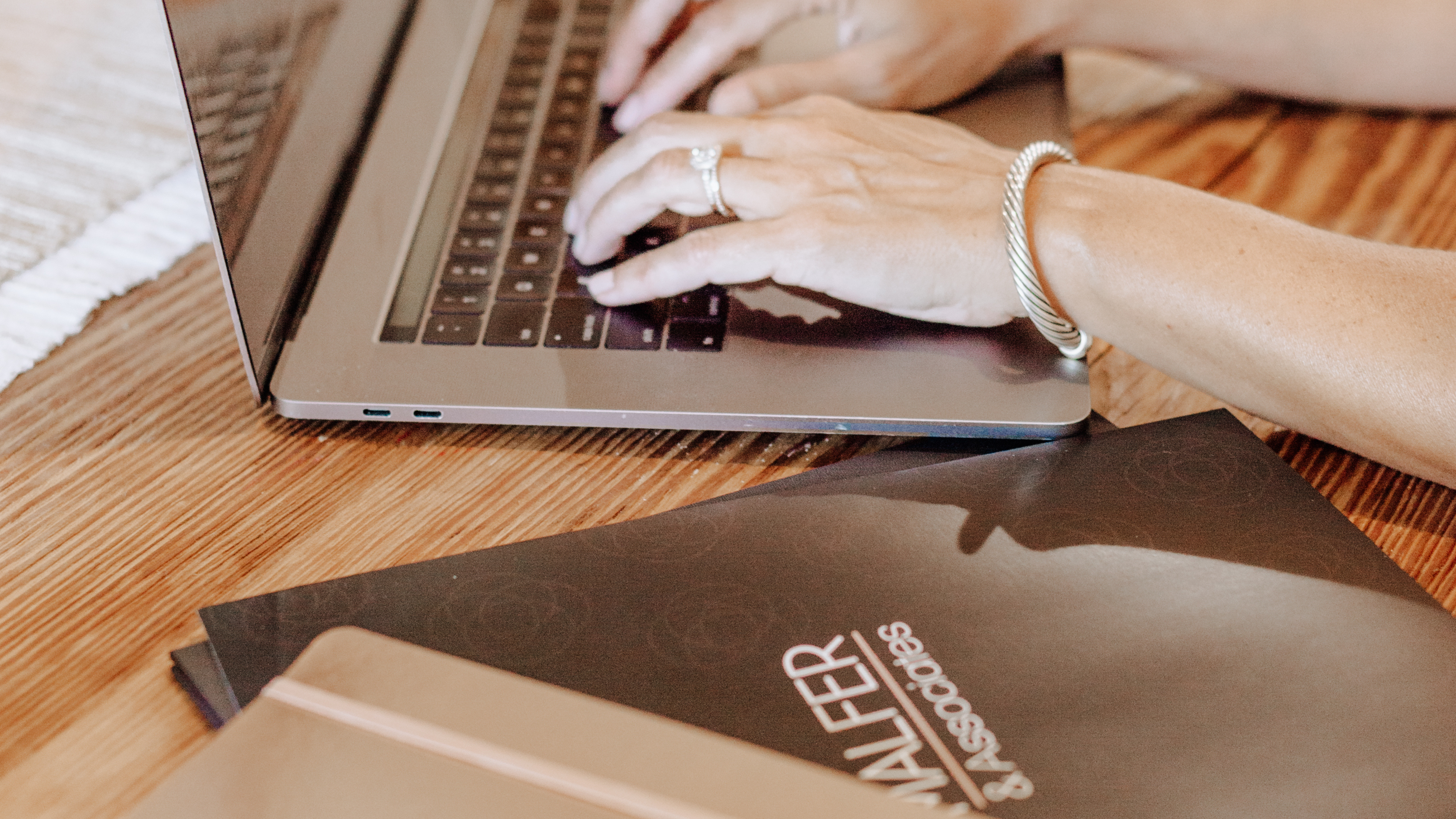 Woman's hands typing on a laptop with a folder of materials and a notebook next to her on the table.